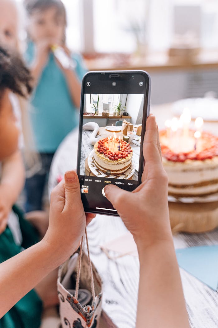 A person photographing a lit birthday cake with a smartphone indoors.